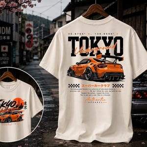May include: An off-white short-sleeve t-shirt featuring a large graphic on the back of an orange sports car with a rear wing. The text reads TOKYO Underground, 35.6764 N 139.6500 E, and Authentic Apparel, along with Japanese characters and checkerboard patterns.