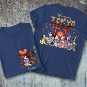 May include: Two navy blue t-shirts with a Disneyland Tokyo theme. One shirt features Mickey Mouse in a kimono with a Japanese pagoda. The other shirt has a graphic of Disney characters in kimonos in front of a Japanese gate.