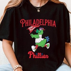 Może przedstawiać: Czarna koszulka z grafiką maskotki Philadelphia Phillies, zielonego stwora z flagą. Tekst "PHILADELPHIA" jest czerwony, z "Phillies" poniżej. Zabawny, sportowy design.