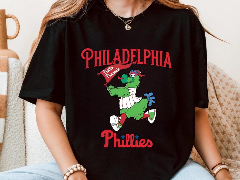 Może przedstawiać: Czarna koszulka z grafiką maskotki Philadelphia Phillies, zielonego stwora z flagą. Tekst "PHILADELPHIA" jest czerwony, z "Phillies" poniżej. Zabawny, sportowy design.