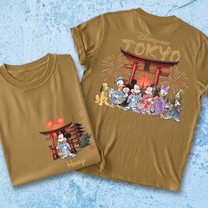 May include: Two tan t-shirts featuring Disney characters in traditional Japanese attire. One shirt shows Mickey Mouse in front of a pagoda, the other displays the characters with the text "Disneyland TOKYO" and a Japanese Torii gate.
