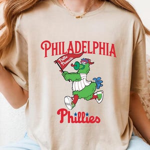 Może przedstawiać: Beżowa koszulka z maskotką Philadelphia Phillies, Phillie Phanatic, trzymającą flagę. Słowo "PHILADELPHIA" jest wygięte u g&oacute;ry na czerwono, z "Phillies" poniżej. Phanatic jest zielony, czerwony i niebieski.