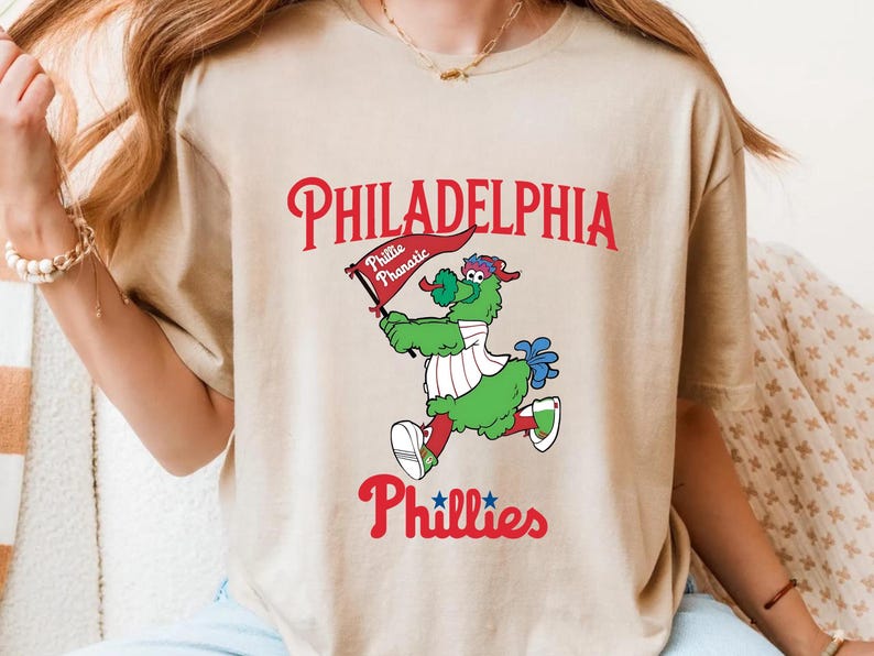 Może przedstawiać: Beżowa koszulka z maskotką Philadelphia Phillies, Phillie Phanatic, trzymającą flagę. Słowo "PHILADELPHIA" jest wygięte u g&oacute;ry na czerwono, z "Phillies" poniżej. Phanatic jest zielony, czerwony i niebieski.