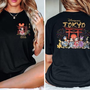 May include: Black t-shirt featuring a front graphic of Mickey Mouse in a kimono, with Japanese text and the name "Olivia." The back displays "Disneyland TOKYO" with Disney characters in traditional Japanese attire, fireworks, and a torii gate.