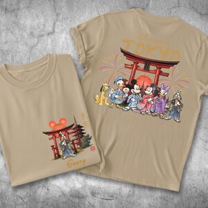 May include: Two tan t-shirts with Japanese-themed Disney character graphics. One shirt features Mickey, Minnie, Donald, Daisy, and Goofy in traditional Japanese attire. The other shirt has a similar design with a pagoda and the word "Goofy".