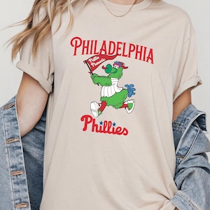 Może przedstawiać: Beżowa koszulka z maskotką Philadelphia Phillies, zielonym stworzeniem trzymającym flagę. Na fladze napis "Phillies Phanatic". Napisy "Philadelphia" i "Phillies" są w kolorze czerwonym.