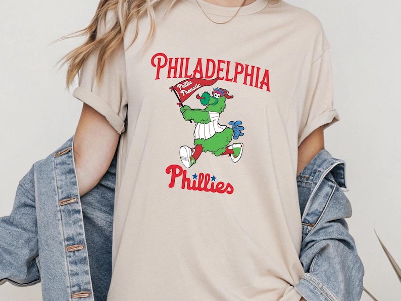 Może przedstawiać: Beżowa koszulka z maskotką Philadelphia Phillies, zielonym stworzeniem trzymającym flagę. Na fladze napis "Phillies Phanatic". Napisy "Philadelphia" i "Phillies" są w kolorze czerwonym.