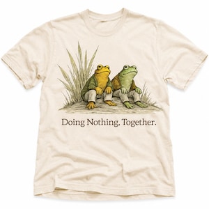 Könnte beinhalten: Ein cremefarbenes Kurzarm-T-Shirt mit einer grafischen Illustration von zwei anthropomorphen Fröschen, die im Gras sitzen. Ein Frosch ist gelb, der andere grün, beide tragen Jacken und Hosen. Der Text lautet: Doing Nothing, Together.