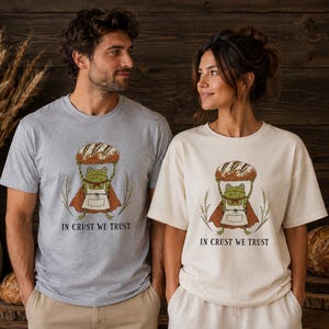 Könnte beinhalten: Ein hellgraues T-Shirt mit Rundhalsausschnitt und ein cremefarbenes T-Shirt mit Rundhalsausschnitt, beide mit einer Grafik eines grünen Frosches in Schürze und Umhang, der ein Brot über dem Kopf hält, mit dem Text "IN CRUST WE TRUST" darunter.