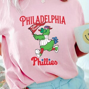 Może przedstawiać: R&oacute;żowa koszulka z napisem "PHILADELPHIA Phillies" w kolorze czerwonym i kresk&oacute;wkową maskotką trzymającą flagę z napisem "Phillie Phanatic". Maskotka jest zielona z białymi i czerwonymi akcentami.