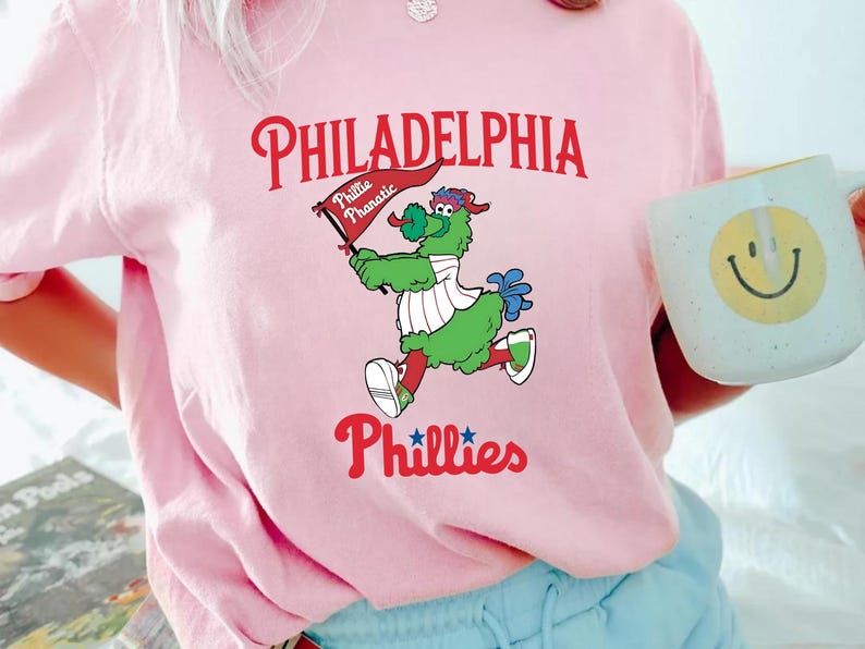 Może przedstawiać: R&oacute;żowa koszulka z napisem "PHILADELPHIA Phillies" w kolorze czerwonym i kresk&oacute;wkową maskotką trzymającą flagę z napisem "Phillie Phanatic". Maskotka jest zielona z białymi i czerwonymi akcentami.