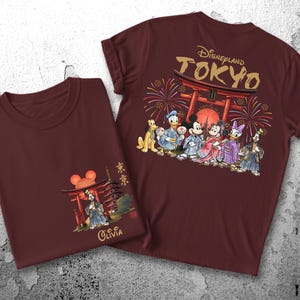 May include: Two maroon t-shirts with Japanese-themed Disney designs. One shirt features a front graphic of a pagoda and Mickey Mouse ears, with the name "Olivia." The other shirt has "Disneyland TOKYO" and Disney characters in traditional Japanese attire.