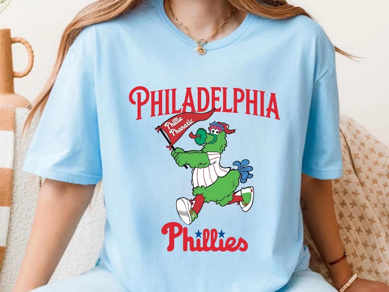 Może przedstawiać: Jasnoniebieska koszulka z napisem "PHILADELPHIA" w kolorze czerwonym, nad kresk&oacute;wkową maskotką trzymającą flagę z napisem "Phillie Phanatic". Słowo "Phillies" znajduje się pod maskotką w kolorze czerwonym.