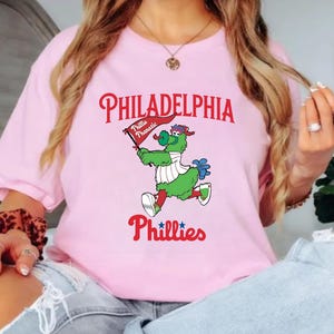 Może przedstawiać: Jasnor&oacute;żowa koszulka z napisem "PHILADELPHIA" w czerwonym skrypcie nad kresk&oacute;wkową maskotką trzymającą flagę z napisem "Phillie Phanatic". Słowo "Phillies" znajduje się poniżej w czerwonym skrypcie.