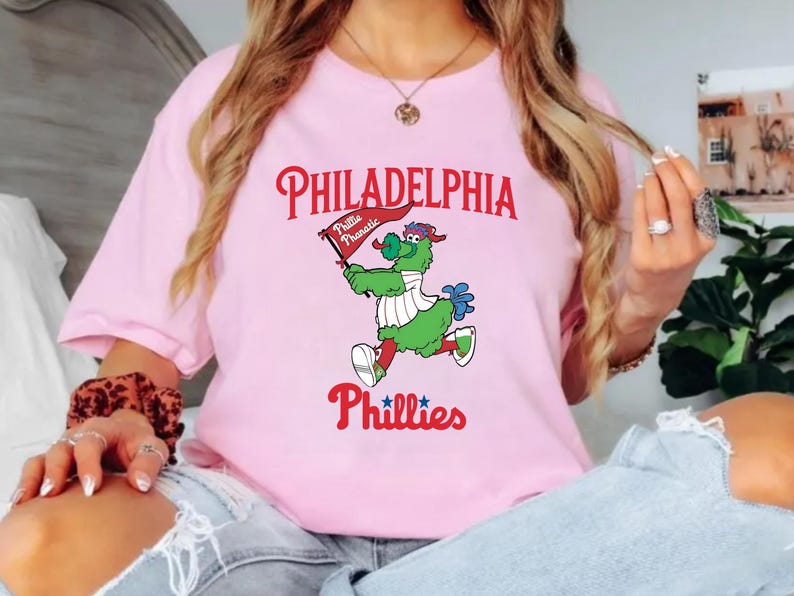 Może przedstawiać: Jasnor&oacute;żowa koszulka z napisem "PHILADELPHIA" w czerwonym skrypcie nad kresk&oacute;wkową maskotką trzymającą flagę z napisem "Phillie Phanatic". Słowo "Phillies" znajduje się poniżej w czerwonym skrypcie.