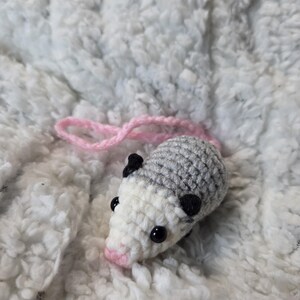 May include: A handmade crochet opossum toy. The opossum has a gray body, white face, and pink nose. It features black eyes and black spots on its head. A pink yarn tail extends from the back. The toy is set against a white, textured background.