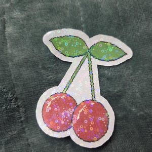 May include: A holographic sticker featuring a pixel art design of two red cherries with green leaves. The sticker has a white border and a shimmering, iridescent finish that reflects light across the surface of the fruit and the leaves.