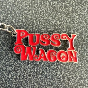 May include: A red and silver keychain with the words "Pussy Wagon" in a decorative font. The keychain is made of metal and has a silver chain attached.