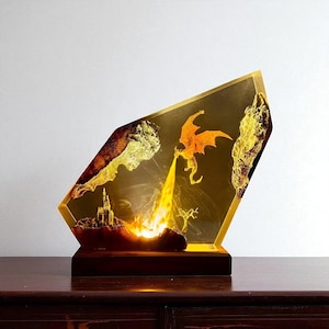 Puede incluir: Una pieza de arte decorativa de resina que representa un dragón que escupe fuego sobre un castillo. La obra de arte está encerrada en un marco angular con bordes dorados, sobre una base de madera oscura. El dragón es naranja y el fuego es amarillo y naranja.
