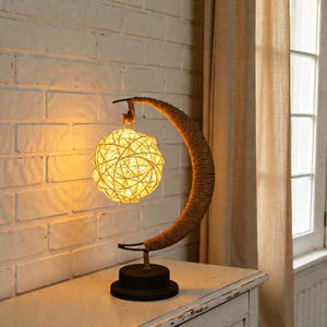 Pode incluir: Uma luminária de mesa decorativa com um braço em forma de crescente envolto em corda e uma luminária esférica tecida. A lâmpada tem uma base preta e emite um brilho quente e convidativo. A lâmpada tem aproximadamente 30 cm de altura.