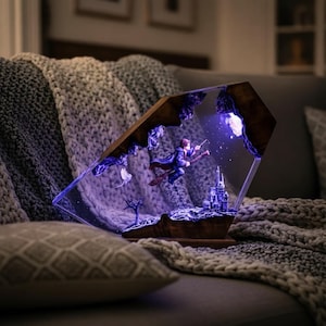 Può includere: Lampada decorativa raffigurante un mago su una scopa volante verso un castello. La lampada ha una cornice in legno e un frontale trasparente e angolato. La scena è illuminata con luci viola e bianche, creando un'atmosfera magica. La lampada è posizionata su una base di legno.