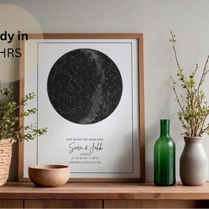 May include: A framed print featuring a black and white star map in a circular design. The print includes the text "THE NIGHT WE WERE MET" and personalized details. A wooden bowl, green bottle, and vase with greenery are on a wooden surface.