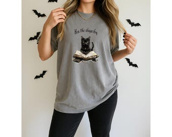 May include: Gray t-shirt featuring a black cat sitting on an open book with the text "Hex the oligarchy." The shirt has a relaxed fit and short sleeves. Black bat decorations are on the wall behind the shirt.