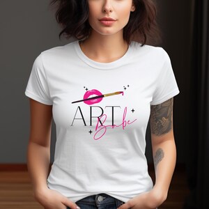 May include: White t-shirt featuring the text "ART Babe" in black and pink script. The design includes a pink lip print, a paintbrush, and small star accents. The shirt has a crew neck and short sleeves.