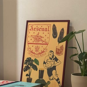 Könnte beinhalten: Gerahmter Druck mit dem Arsenal-Logo und Illustrationen. Das Kunstwerk zeigt einen Rugby-Spieler, Schuhe und einen Cocktail. Der Druck hat einen weinroten Rahmen und einen cremefarbenen Hintergrund mit roten und schwarzen Illustrationen. Das Wort "Arsenal" steht oben.