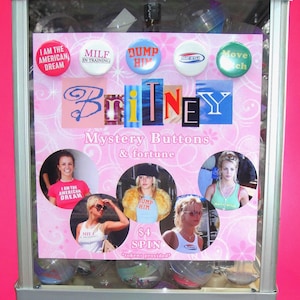 May include: A vintage gumball machine with a pink and white exterior. The machine dispenses "Mystery Buttons & fortune". The buttons feature various phrases and images. The machine has a coin slot and a dispensing area.