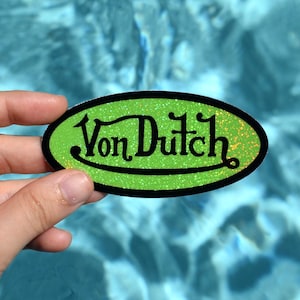 May include: An oval-shaped sticker with the text "Von Dutch" in black script. The sticker has a green glitter background and a black border. The sticker is held against a blue water background.