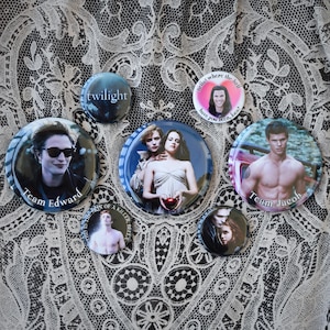 May include: A collection of round buttons featuring images and text from the Twilight saga. The buttons display characters, phrases like "Team Edward" and "Team Jacob", and the word "Twilight". The buttons are arranged on a patterned, lacy surface.