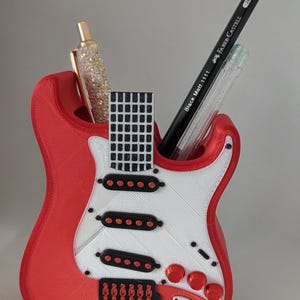 Puede incluir: Un organizador de escritorio rojo con forma de guitarra y un golpeador blanco. El organizador contiene un bolígrafo dorado, un lápiz negro con "Faber-Castell" impreso y un bolígrafo transparente. Este organizador es un artículo único y funcional.