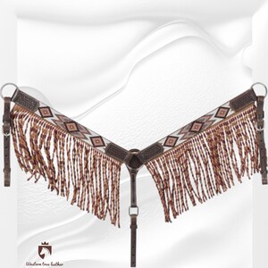 May include: A brown leather horse breast collar featuring a V-shaped design with a white and brown geometric diamond pattern inlay. The piece includes long, braided brown leather fringe hanging from the bottom and silver-toned metal hardware.