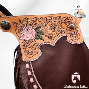 May include: A pair of dark brown leather chaps featuring a tan leather yoke with intricate floral tooling. The roses are hand-painted in soft pink with green leaves. The chaps include long fringe, silver-toned metal conchos, and a woven leather trim.