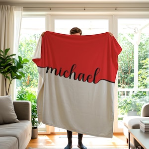 May include: A personalized blanket with a red and white color block design. The word "michael" is written in a black script font across the blanket. The blanket is being held up in front of a window.