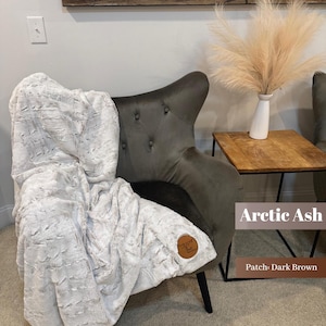 May include: A plush, light gray blanket draped over a dark gray armchair. The blanket has a textured pattern and a brown leather patch with a logo. The words "Arctic Ash" and "Patch: Dark Brown" are visible.