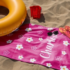 Puede incluir: Una toalla de playa rosa brillante con estampados de margaritas blancas y el nombre "Charlotte" en escritura blanca. La escena incluye un flotador inflable amarillo, un cubo rojo, gafas de sol y chanclas naranjas sobre la arena.