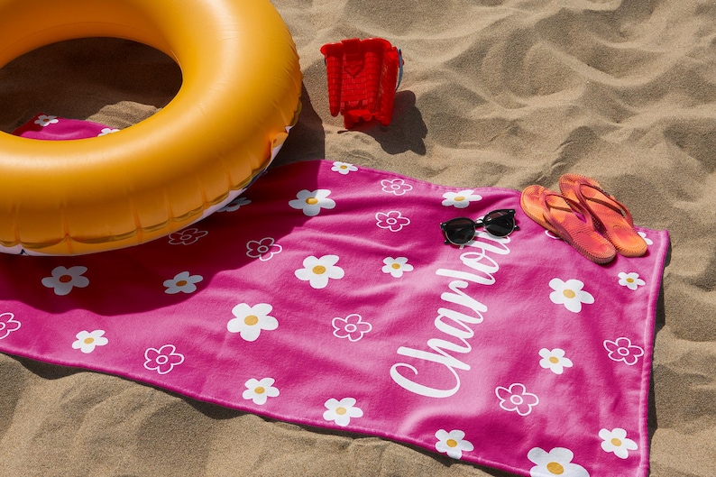 Puede incluir: Una toalla de playa rosa brillante con estampados de margaritas blancas y el nombre "Charlotte" en escritura blanca. La escena incluye un flotador inflable amarillo, un cubo rojo, gafas de sol y chanclas naranjas sobre la arena.