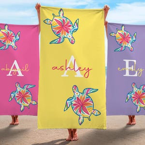 May include: Three beach towels in pink, yellow, and purple, each featuring a colorful sea turtle design. The towels have personalized names in white script: Abigail, Ashley, and Emily. The towels are held up on a beach.