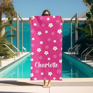 Puede incluir: Una toalla de playa rosa brillante con un patr&oacute;n floral repetido de flores blancas y amarillas. El nombre "Charlotte" est&aacute; escrito en blanco. La toalla se sostiene frente a una piscina.