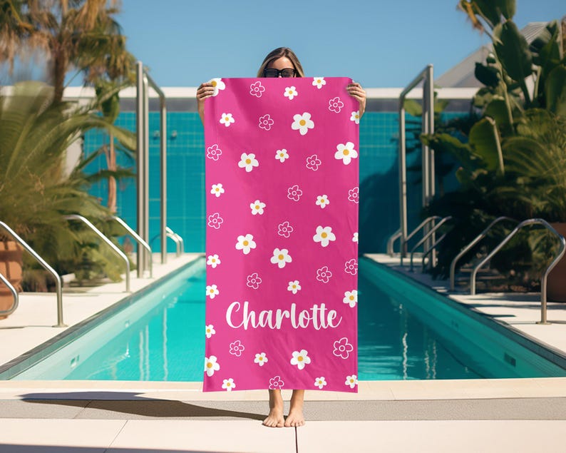 Puede incluir: Una toalla de playa rosa brillante con un patr&oacute;n floral repetido de flores blancas y amarillas. El nombre "Charlotte" est&aacute; escrito en blanco. La toalla se sostiene frente a una piscina.