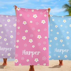 Puede incluir: Tres toallas de playa en tonos lavanda, rosa y azul, cada una adornada con un patr&oacute;n de flores blancas. El nombre "Harper" est&aacute; impreso en escritura blanca en cada toalla. Las toallas se sostienen en una playa.