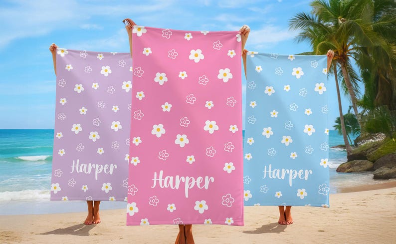 Puede incluir: Tres toallas de playa en tonos lavanda, rosa y azul, cada una adornada con un patr&oacute;n de flores blancas. El nombre "Harper" est&aacute; impreso en escritura blanca en cada toalla. Las toallas se sostienen en una playa.