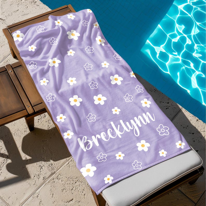 Puede incluir: Una toalla de playa lavanda con un estampado de margaritas blancas y el nombre "Brecklynn" en escritura blanca. La toalla est&aacute; colocada sobre una tumbona de madera junto a una piscina.
