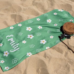 Puede incluir: Una toalla de playa verde con un estampado de margaritas blancas y el nombre "Emily" en escritura blanca. Una bolsa redonda tejida y gafas de sol descansan sobre la toalla, que est&aacute; extendida sobre arena.