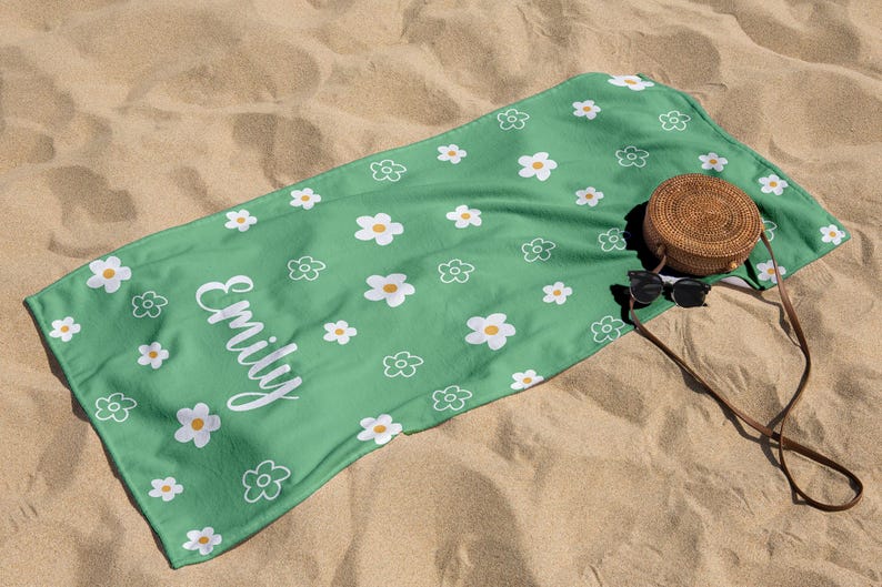 Puede incluir: Una toalla de playa verde con un estampado de margaritas blancas y el nombre "Emily" en escritura blanca. Una bolsa redonda tejida y gafas de sol descansan sobre la toalla, que est&aacute; extendida sobre arena.