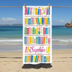 May include: White beach towel with a colorful bookshelf design. The towel features rows of books in various colors, with floral and leaf accents. The name "Sophia" is written in pink script. The towel is hanging on a clothesline at the beach.