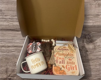 May include: A gift box with fall-themed items. Includes a white enamel mug with "Hello Pumpkin" text, a wooden sign with a pumpkin design, a small broom, and a decorative pumpkin. The box is lined with red and brown filler.