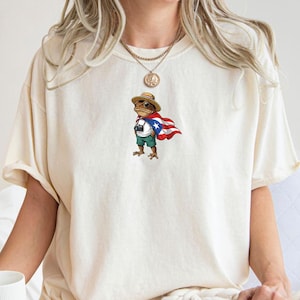 Könnte beinhalten: Cremefarbenes T-Shirt mit einer Cartoon-Froschfigur, die einen Strohhut, Shorts und einen Umhang mit dem Design der puerto-ricanischen Flagge trägt. Der Frosch hält eine Kamera. Das Shirt hat kurze Ärmel und eine lockere Passform.
